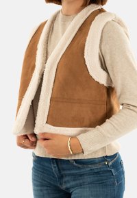 Brun faux suede väst med vit sherpa-foder, ärmlös design, två framfickor och en mjuk textur. Bärs över en ljus tröja.