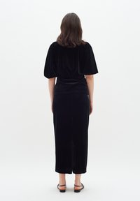 Completo in velluto nero formato da due pezzi, composto da una blusa ampia con maniche a sbuffo e una gonna aderente lungo la caviglia. Texture liscia e design minimalista.