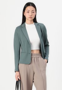 Teal bleiser piiratud disainiga, kahe esiosa taskuga ja ühe nupuga kinnitusega, koos heleda beeži vabaltistuva pükstega ja valge lühikese topiga.