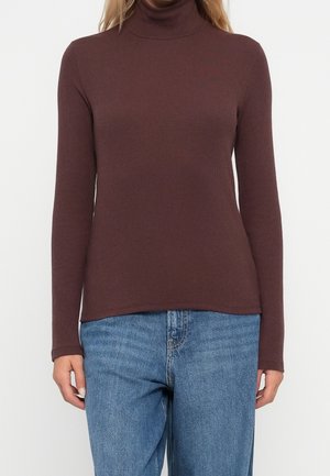 Nainen pukeutuneena istuvaan pitkähihainen ruskea pooloneule ja väljät siniset farkut, seisoo yksinkertaisen valkoisen taustan edessä.