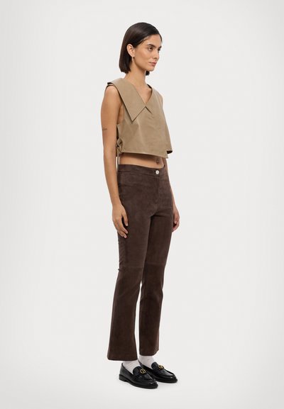 Beiger Cropped-Top mit großem Kragen und seitlicher Raffung, kombiniert mit braunen Wildlederhosen im Schlag und glänzenden schwarzen Loafers mit goldenen Akzenten.