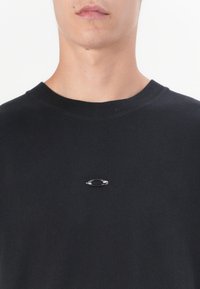 Jeune personne portant un t-shirt noir à col rond avec un petit logo ovale métallique centré sur la poitrine.