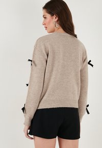 Un pull en tricot beige à manches longues, avec des accents de ruban noir sur les poignets, un ourlet côtelé, et une coupe décontractée. Des shorts noirs sont portés en dessous.