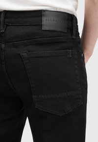 Vaqueros de mezclilla negra con una textura suave, que presentan una etiqueta de cuero negro en la cintura y bolsillos traseros diagonales. Detalles de diseño simples.
