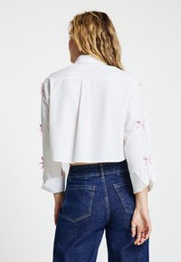 White cropped shirt with long sleeves, featuring pink ribbon accents on the sleeves. Paired with high-waisted blue denim pants.