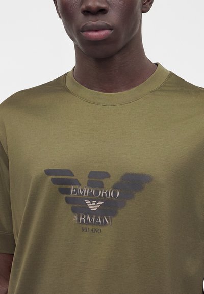 Частичен изглед на човек, носещ тениска Emporio Armani Milano в маслиненозелен цвят с черно лого с криле на гърдите.