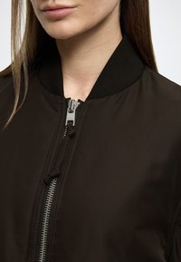 Svart bomberjakke med jevn tekstur, ribbet krage og en fremtredende glidelåsdetalj foran, lett åpnet.