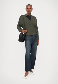 Blazer de doble botonadura en verde oliva, top negro, jeans de pierna ancha en azul oscuro, bailarinas blancas de punta y un bolso tejido negro texturizado.