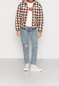 Giubbotto imbottito a quadri marroni e bianchi con il testo "KARL KANI", indossato sopra un felpa crema, abbinato a jeans blu strappati e sneakers beige.