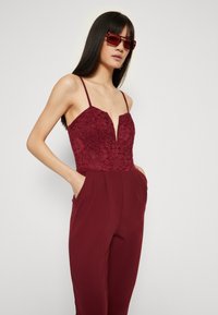 Burgunderfarget jumpsuit med blondetopp, dyp V-hals, tynne stropper og tapered ben. Den har sidelommer og en jevn, strukturert finish.