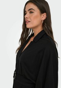 Crna košulja s ovratnikom i dugim rukavima, koja ima strukiran pojas. Tkanina izgleda glatko s oblikovanim krojem i elegantno pada.