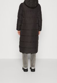 Black puffer coat with a hood, long length, quilted design, and horizontal stitching. Worn over gray pants and black platform boots.