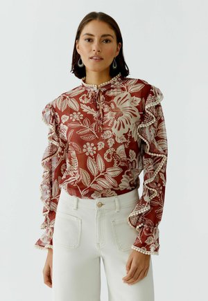 Woman wearing a rust-red floral patterned blouse with ruffled sleeves and white high-waisted pants, standing against a plain light background.