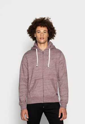 Sudadera con cremallera de color marrón burdeos, con una tela texturizada, cordones blancos, bolsillos frontales y puños acanalados. Combinada con pantalones negros.