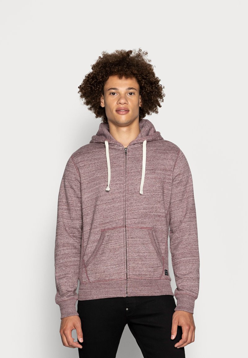 Sweatshirt zippé bordeaux en tissu texturé, avec des cordons blancs, poches frontales et poignets côtelés. Associé à un pantalon noir.