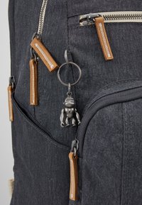 Sac à dos en tissu gris foncé avec plusieurs curseurs de fermeture éclair beige et un petit porte-clés robot en argent suspendu à une fermeture éclair.