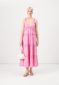 Pink maxi dress with ruffled tiers, deep V-neck, and thin straps. Paired with strappy beige sandals and a small white handbag.