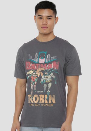 Junger Mann trägt ein dunkelgraues Batman- und Robin-Grafik-T-Shirt mit Vintage-Stadtbild und Text auf der Vorderseite.