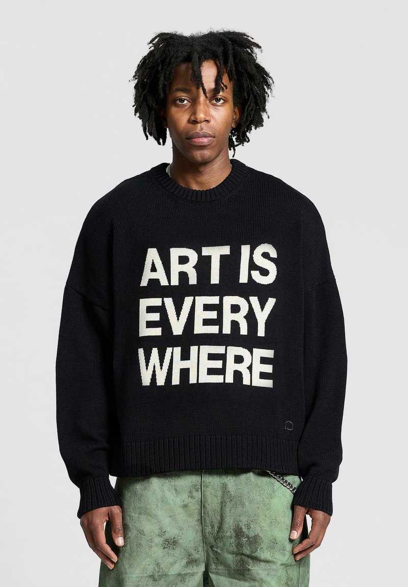 Maglione nero lavorato a maglia con la scritta "L'ARTE È OVUNQUE" in crema. Presenta un colletto rotondo e polsini a coste. Abbinato a pantaloni verde texturizzati.