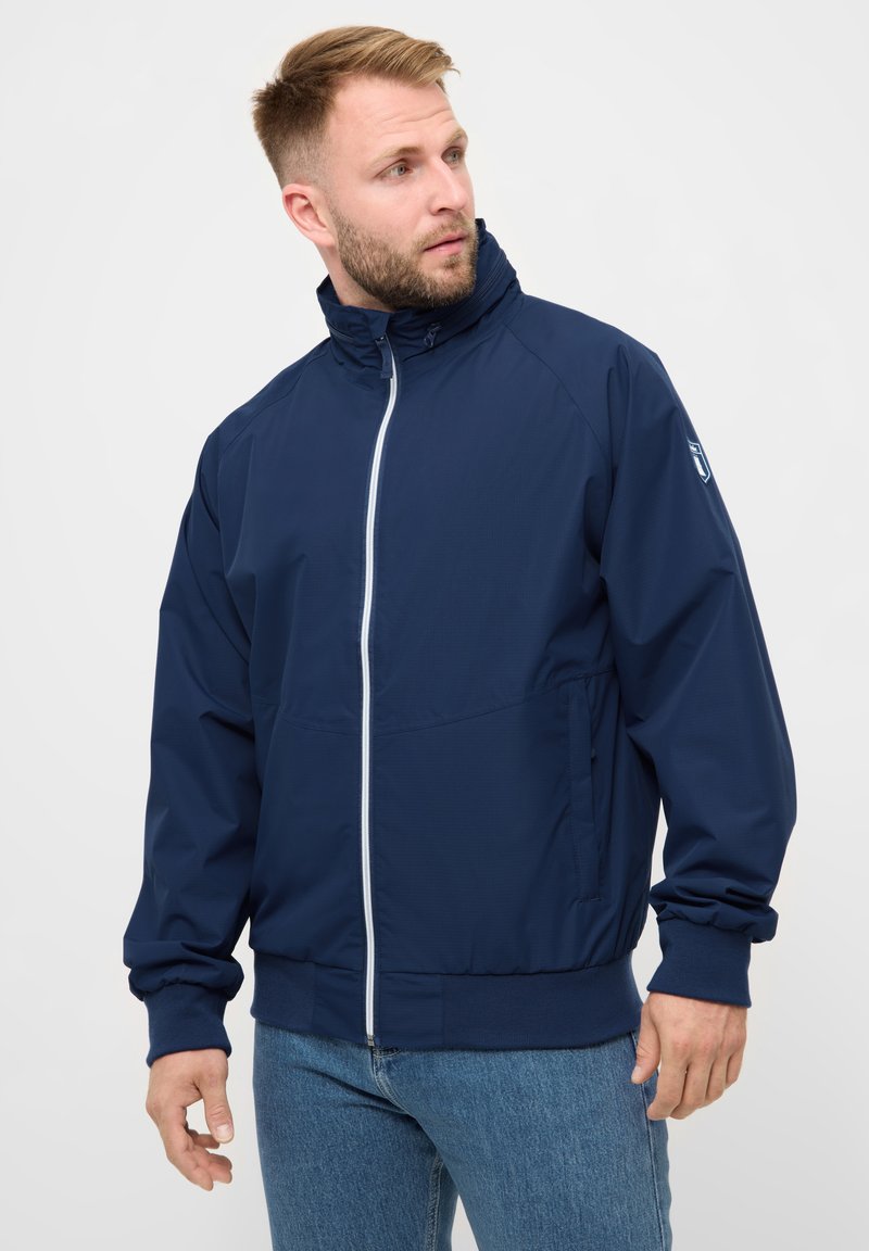 Mann trägt eine marineblaue Reißverschlussjacke und blaue Jeans und schaut auf einem schlichten Hintergrund nach links.
