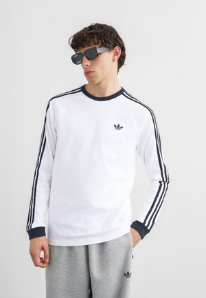 Mladý muž v bílé dlouhém tričku Adidas s námořnicky modrými pruhy, šedých teplácích Adidas, černých slunečních brýlích a zlatém náušnici, stojící u bílé zdi.