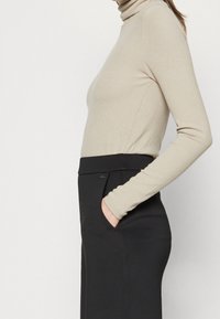 Donna che indossa un maglione beige a collo alto e una gonna nera a vita alta, con una mano in tasca, mostrata di lato dalla vita in su.