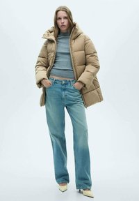 Beige pufferjack met een capuchon, grijs cropped sweater en lichtblauwe spijkerbroek. Het model staat met de handen in de zakken en draagt puntige witte schoenen.