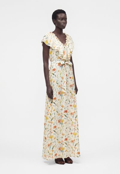 Γυναίκα που φοράει λευκό μακρύ φόρεμα χωρίς μανίκια με ραμμένο ντεκολτέ και πορτοκαλί, κίτρινο ανθικό σχέδιο, στέκεται μπροστά σε απλό φόντο.