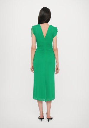 Frau mit langen dunklen Haaren, die ein grünes Midikleid mit kurzen Ärmeln und offenem Rücken trägt, steht mit dem Rücken zur Kamera, schwarze Absatzschuhe an.