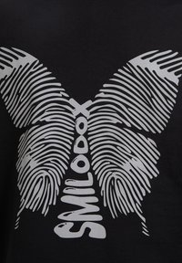 Sort t-shirt med et stort sølvfarvet sommerfugleprint lavet af fingeraftryk mønstre, med ordet "SMILODOX" centreret nedenfor.