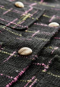Tessuto misto lana di colore nero con un motivo a quadri colorato, caratterizzato da righe rosa e verdi. Bottoni a forma di conchiglia in oro aggiungono un dettaglio.