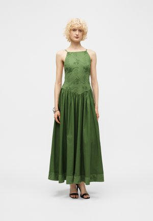 Γυναίκα με ξανθά σγουρά μαλλιά που φοράει αμάνικο πράσινο μακρύ φόρεμα με κεντημένες λεπτομέρειες και μαύρα ανοιχτά ψηλοτάκουνα, στέκεται μπροστά σε απλό φόντο.