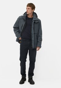 Donkerblauwe, geïsoleerde jas met een hoge kraag en meerdere ritszakken, in combinatie met donkere broek en zwarte laarzen.