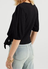 Czarna top z teksturowanym wykończeniem, z puffymi rękawami, dopasowaną talią i detalem w postaci wstążki z tyłu. Zestawiony z jasnoniebieskimi jeansami o wysokim stanie.