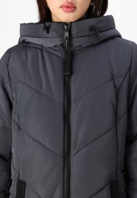 Veste rembourrée grise avec un motif en chevrons, fermeture éclair, capuche réglable et accents noirs sur les poches. Texture douce et lisse.