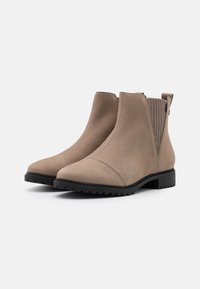 Par de botas de tobillo beige con suelas de goma negra y paneles laterales elásticos, mostradas sobre un fondo blanco.
