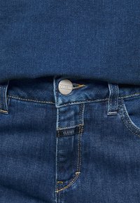 Gros plan sur un jean en denim bleu montrant un bouton en métal et une petite étiquette en tissu, tous deux embossés du mot "CLOSED."