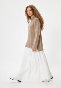 Maglione beige lavorato a maglia con collo alto, maniche lunghe e polsini a coste, abbinato a una gonna bianca fluida e scarpe bianche a punta.