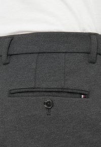 Sötétszürke öltönynadrág hátsó része, gombos zsebével és egy kis piros, fehér és kék szövet címkével a zseb széle közelében.