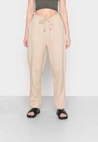 Pantalon en lin beige avec une coupe décontractée, taille élastique, cordon de serrage et deux poches latérales. Associé à des sandales plates noires.
