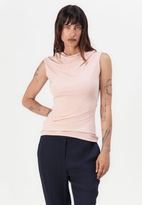 Lys rosa ermeløs topp med cowl-hals, laget av et glatt stoff, sammen med mørk marineblå bukser. Har arm tatoveringer.