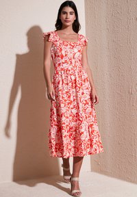 Punainen kukallinen midi-mekko, jossa on frilla-hihnat, neliön muotoinen pääntie, kiristetty vyötärö ja kerroksellinen hame. Yhdistetty beigeihin nilkkapanta-sandaaleihin.
