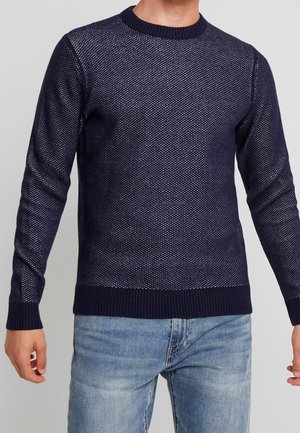 Homme portant un pull en maille texturée bleu marine avec poignets et ourlet côtelés, associé à un jean en denim bleu clair.