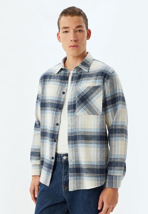Camisa de franela con un patrón a cuadros en azul y crema, cierre de botones, un bolsillo en el pecho y textura suave con mangas largas.