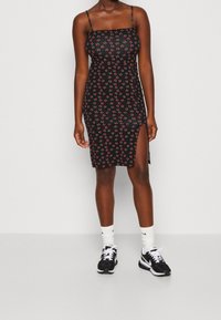 Schwarzes, figurschmeichelndes Kleid mit dünnen Trägern, das mit einem roten und grünen Kirschmuster bedruckt ist. Das Kleid hat einen seitlichen Schlitz und wird mit schwarzen Sneakers und weißen Socken kombiniert.