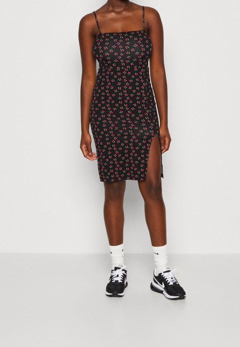 Schwarzes, figurschmeichelndes Kleid mit dünnen Trägern, das mit einem roten und grünen Kirschmuster bedruckt ist. Das Kleid hat einen seitlichen Schlitz und wird mit schwarzen Sneakers und weißen Socken kombiniert.