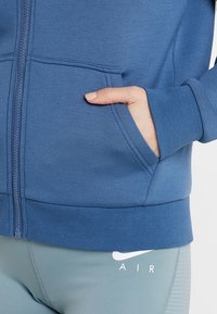 Main dans la poche d'une veste bleue à fermeture éclair portée sur un legging Nike Air bleu clair sur fond blanc.