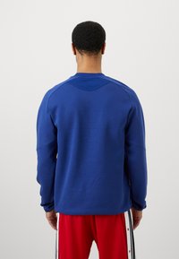 Plavi dukserica s teksturiranim panelom na ramenima, dugim rukavima i okruglim izrezom, u kombinaciji s crvenim hlačama koje imaju bočne gumbe.