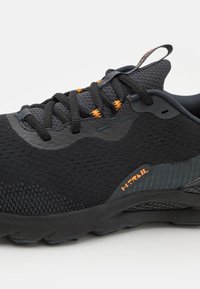 Chaussure de sport noire avec une tige en mesh texturé et des accents gris, présentant des détails orange sur les lacets et le logo sur le côté, ainsi qu'une semelle à motifs.