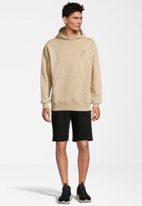 Felpa beige con cappuccio e tasca frontale, logo sottile, abbinata a pantaloni corti neri e sneakers nere, in piedi su uno sfondo neutro.
